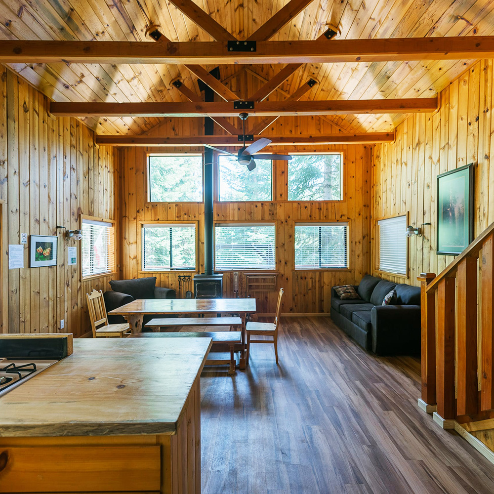 Spacious open-concept cabin living and dining area with vaulted wood-paneled ceilings, exposed beams, a wooden dining table, a black sofa, a wood-burning stove, and multiple large windows.