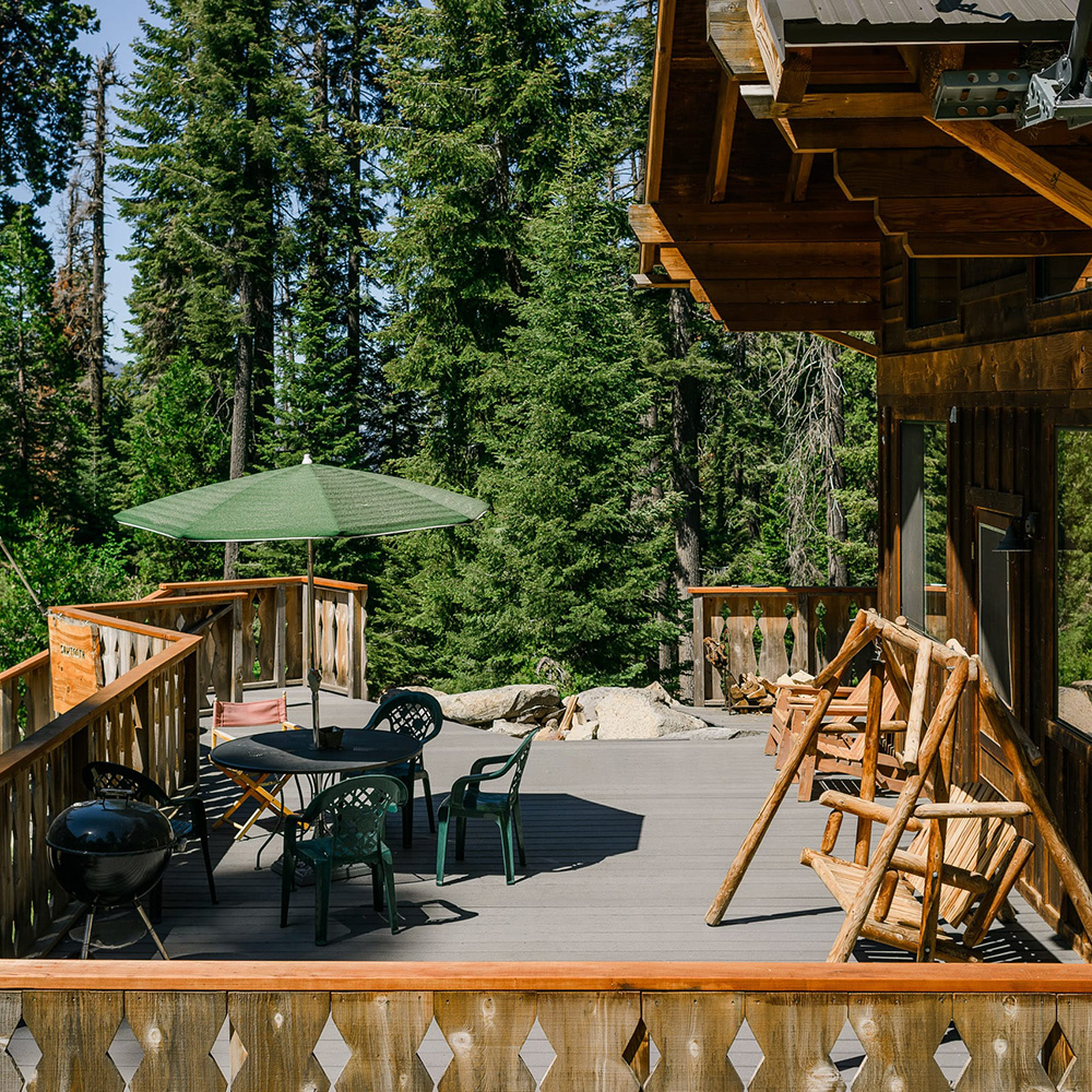 Wooden cabin deck surrounded by pine trees, equipped with a patio table and green umbrella, a black charcoal grill, and a rustic wooden swing bench.