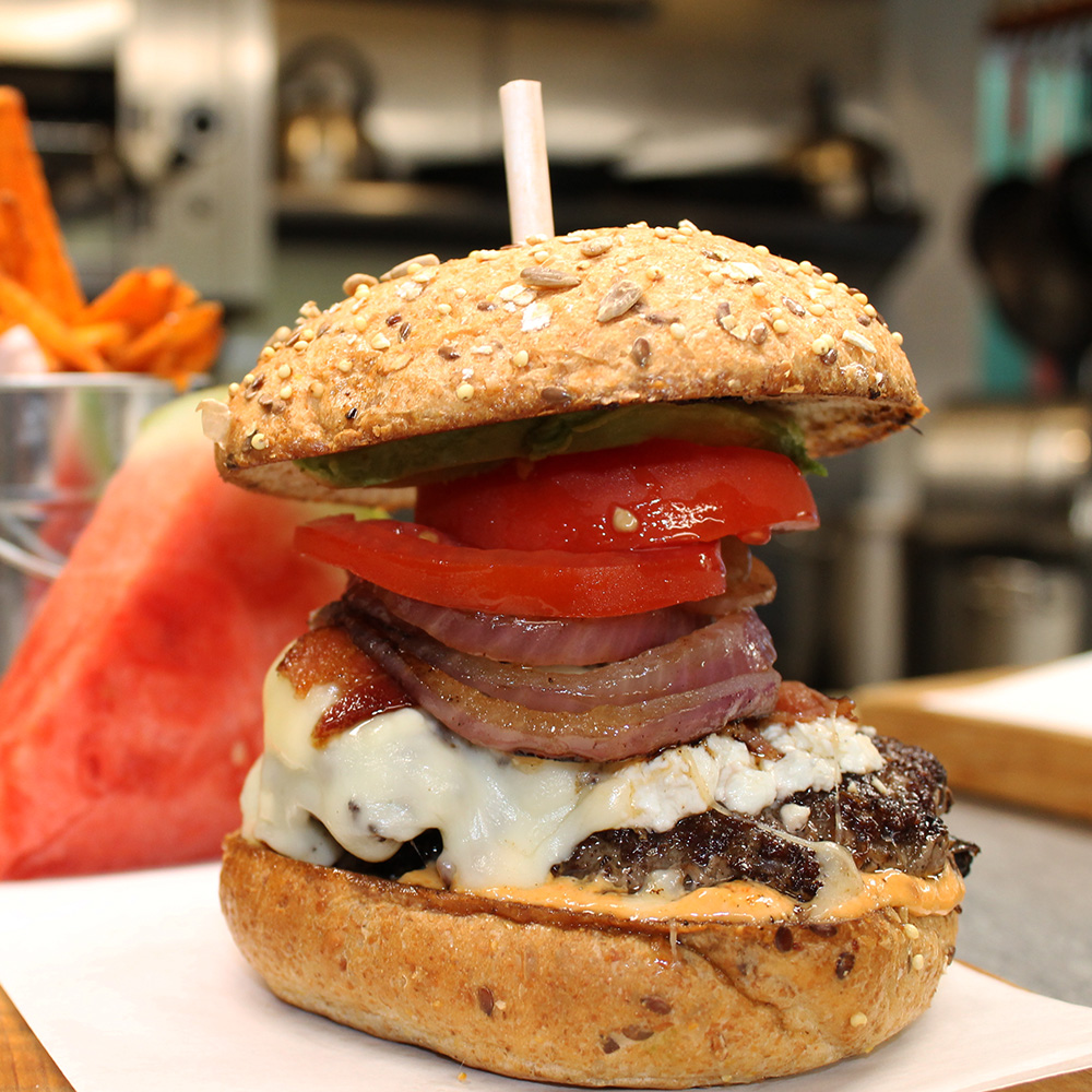 A tall gourmet burger on a seeded bun featuring a beef patty, melted cheese, bacon, red onion, thick tomato slices, avocado, and sauce, with sweet potato fries and a watermelon slice in the background.