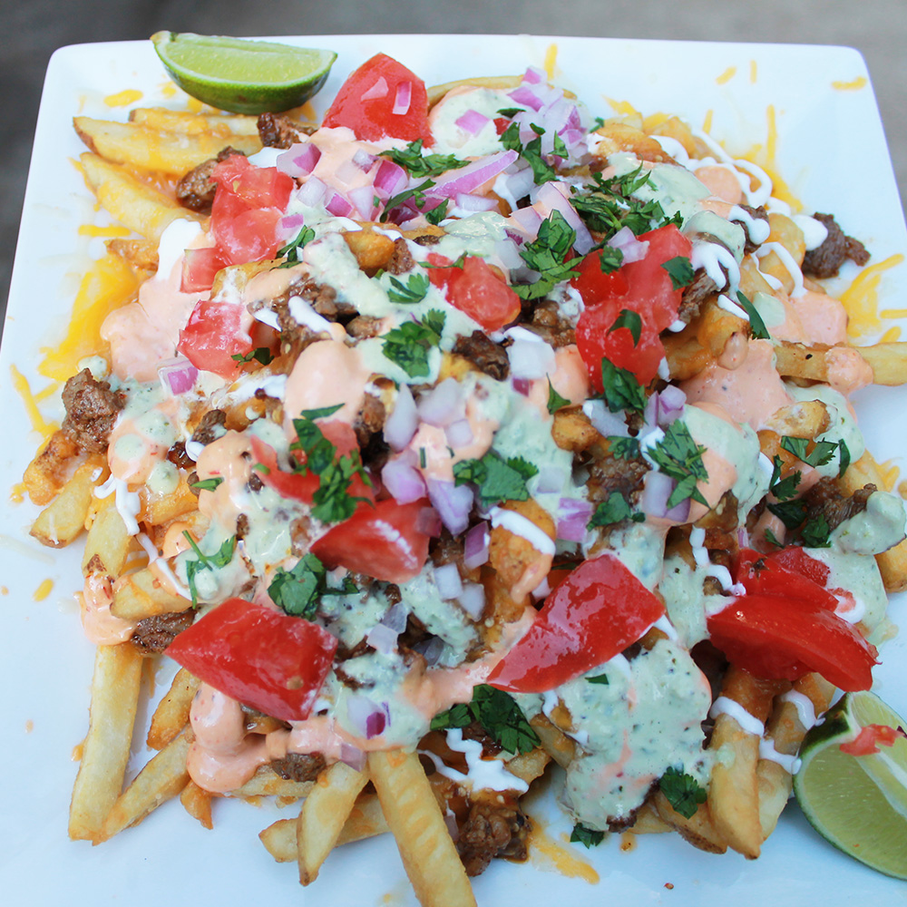 A square plate of loaded french fries topped with melted cheese, ground meat, diced tomatoes, red onion, cilantro, and drizzled with multiple creamy sauces, garnished with lime wedges.