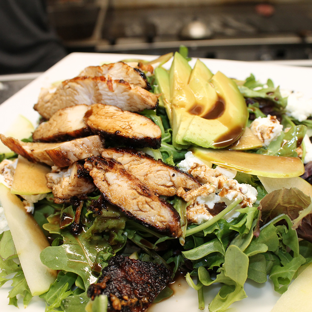 A close-up of a mixed green salad topped with sliced grilled chicken breast, fresh avocado, sliced pears or apples, white cheese crumbles, and a dark balsamic drizzle.